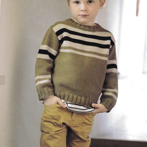 May include: A child's green sweater with black and white horizontal stripes, paired with mustard-coloured trousers. The sweater features a ribbed neckline and cuffs. The child has their hands in their pockets. The outfit is casual.
