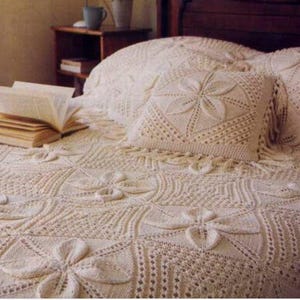 May include: Cream-colored, knitted bedspread with a floral pattern. A matching pillow with a large flower design sits on the bed. An open book rests on the bedspread. The bed is in a bedroom with a wooden headboard.