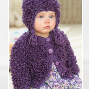 May include: A child wearing a matching purple knitted hat and cardigan. The hat has ear flaps and tassels, and the cardigan has button closures. The outfit is made of a textured, looped yarn. The child is also wearing a floral print dress.