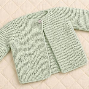 May include: A pale green knitted baby cardigan with long sleeves and a single button fastening. The cardigan is made from a soft, textured yarn and has a simple, classic design. The sleeves have folded cuffs.