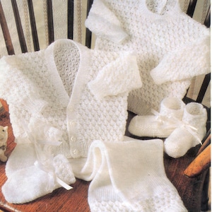 May include: A set of white knitted baby clothes, including a cardigan, sweater, leggings, and booties. The garments feature a textured pattern and are arranged on a wooden chair. The booties are tied with white ribbons.