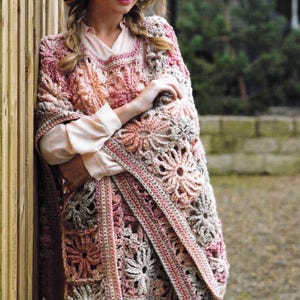May include: A handmade crochet shawl in shades of pink, beige, and gray. The shawl features a floral pattern and a fringed edge. The woman in the image is wearing a light-colored shirt and a brown hat.