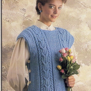 May include: A light blue knitted vest with a cable knit pattern and button closures. The vest is sleeveless and has a scoop neck. The model is wearing a white blouse underneath the vest and is holding a bouquet of pink and yellow roses.