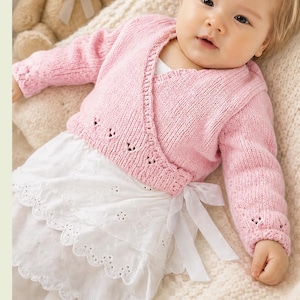May include: A baby wearing a light pink, knitted wrap sweater with long sleeves and decorative eyelet details. The baby is also wearing a white eyelet lace skirt with a white ribbon tie. The baby is lying on a cream-colored blanket.