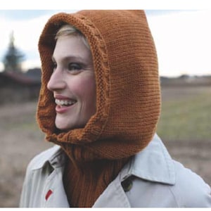May include: A woman wearing a knitted hood in a warm, earthy brown color. The hood is a cowl style, covering the neck and head, with a textured knit pattern. The woman is wearing a light-colored coat.