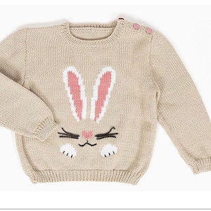 May include: A beige knitted sweater with a bunny face design. The bunny has white ears with pink insides, black whiskers, and a pink nose. The sweater has long sleeves and small pink buttons on the shoulder.