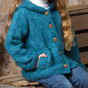 May include: Teal hooded cardigan with wooden buttons and pockets. The knitted jacket has a textured appearance and is worn with blue jeans. The cardigan is a cosy, handmade garment.