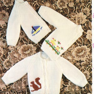 May include: Three off-white knitted baby cardigans are shown, each with button fastenings. One has a sailboat motif, another a floral design, and the third a brown squirrel. They are displayed on a floral patterned background.