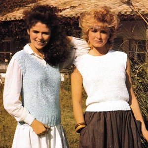 May include: Two women wearing sleeveless knitted tops. The woman on the left is wearing a light blue top and a white shirt underneath. The woman on the right is wearing a white top. Both women are standing in front of a house with a brown roof.