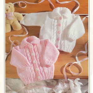 Puede incluir: Dos cárdigans de punto para bebé, uno rosa y otro blanco, con cierre de botones y un patrón de punto de cable. Ambos cárdigans tienen un pequeño diseño de flor en los botones.