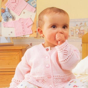 May include: A light pink baby cardigan with a button-up front and decorative openwork detailing on the cuffs and hem. The image also shows a flat lay of the same cardigan design, along with a small doll. The cardigan is made of a soft, knitted material.