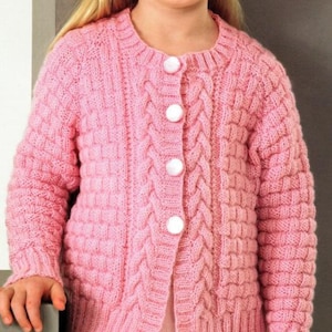 May include: A light pink knitted cardigan with a round neckline and long sleeves. The sweater features a cable knit design down the front and on the sleeves, with four white buttons. The cardigan is a children's garment.