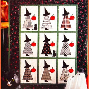 May include: A Halloween-themed quilt featuring nine square panels. Each panel depicts a witch holding a red apple, with a black cat in some panels. The witches wear black hats and patterned dresses. The quilt has a black floral border.