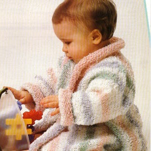 May include: A baby wearing a white, pink, and green striped knitted robe. The robe has a shawl collar and long sleeves. The baby is sitting on a white surface and playing with a toy.