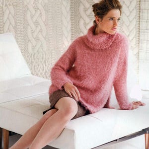 Pode incluir: Um suéter rosa macio, de gola alta e grande. O suéter tem mangas compridas e uma textura fofa. A modelo está usando uma saia marrom. O fundo apresenta um banco branco e uma parede estampada.