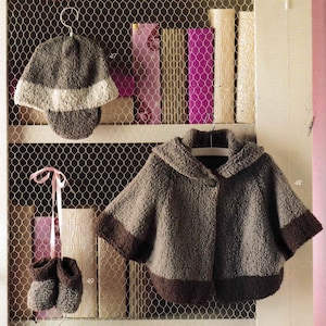 Può includere: Esposizione di abbigliamento per bambini fatto a mano, tra cui un cappello grigio e bianco, un mantello con cappuccio con bordi marroni e scarpine abbinate. Gli articoli sono esposti su una mensola di filo metallico con libri sullo sfondo.