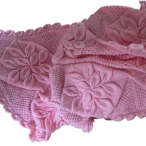 May include: A pale pink knitted blanket with a floral design. The blanket has raised, three-dimensional flower patterns and a scalloped edge. The texture appears delicate and cosy, suitable for a baby or as a decorative throw.