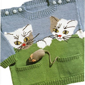 May include: A light blue knitted sweater with a green pocket and a mouse toy. The sweater features two white and brown cat faces peeking over a green field. The sweater has three light blue buttons on the shoulder.