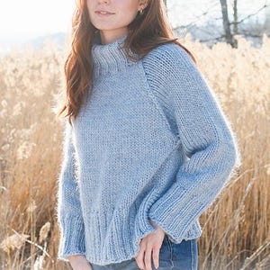 May include: A pale blue knitted jumper with a high, ribbed neckline and long sleeves. The jumper has a textured finish and a slightly flared hem. The model is wearing blue jeans. The background features tall, dry grass.