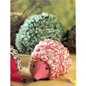 May include: A handmade plush hedgehog with a pink body and a head of red and white yarn. The hedgehog has black bead eyes and a black nose. A green pom-pom and a red hedgehog are in the background.