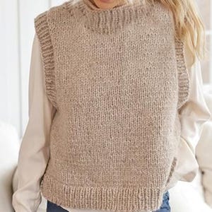 May include: A beige, knitted vest with a high neckline. The sleeveless garment is made of a thick, textured yarn. The vest has a ribbed hem and neckline. The model is wearing a white shirt underneath the vest.