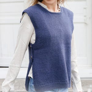 May include: A blue knitted sleeveless sweater vest with a crew neck and buttoned side. It is worn over a white long-sleeved shirt. The texture of the vest is visible, and the buttons are a contrasting colour.
