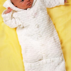 May include: A baby in a white knitted hooded sleepsuit with button fastenings. The sleepsuit has a textured knit pattern and a pocket detail. The baby is lying on a yellow surface.