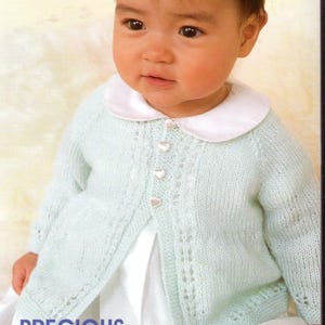 Op de afbeelding: Een baby draagt een lichtblauw gebreid vestje met een witte kraag en hartvormige knopen. Het vestje heeft een delicaat kanten ontwerp. Het woord "PRECIOUS" staat onderaan de afbeelding.