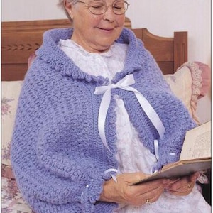 Peut inclure: Un châle en crochet bleu pervenche avec un col à volants et des liens en ruban de satin blanc. Le châle est porté sur une robe blanche. La personne porte des lunettes et lit un livre.
