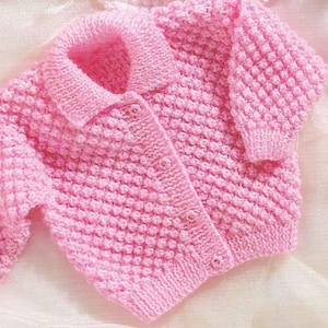 May include: A soft pink, hand-knitted baby cardigan with a textured, bobble stitch pattern. The sweater features a collared neckline, long sleeves, and button closures. A cozy and charming garment for a baby.