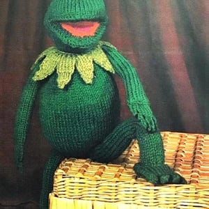 Puede incluir: Una muñeca de rana verde tejida a mano con ojos blancos y negros, boca roja y cuello verde. La rana está sentada en una cesta de mimbre. Los brazos y las piernas de la rana están extendidos.