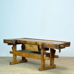 Könnte beinhalten: Eine große, antike Werkbank aus Holz mit natürlicher Oberfläche. Die Werkbank hat eine Schublade, einen Schraubstock und ein unteres Regal. Die Beine und der Rahmen sind stabil, und das Holz weist Gebrauchsspuren auf. Die Werkbank ist ca. 183 cm lang.