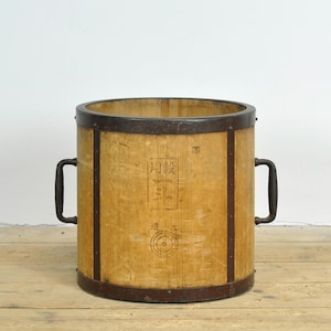 Könnte beinhalten: Ein zylindrischer Holzeimer mit dunklen Metallbändern und zwei seitlichen Griffen. Das helle Holz weist eine vertikale Maserung sowie dunkle japanische Schriftzeichen und kreisförmige Markierungen auf, die Altersspuren zeigen.