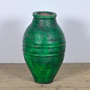 Pode incluir: Um vaso de cerâmica verde, de estilo antigo, com um corpo largo e um pescoço estreito. O vaso tem uma superfície texturizada e ranhuras horizontais ao redor do corpo. A cor verde é irregular, com manchas mais escuras.