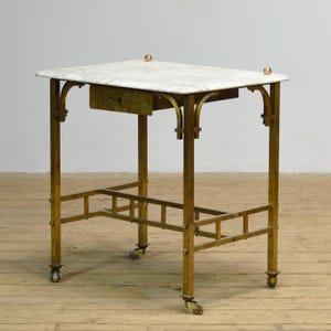 Può includere: Un tavolino antico con piano in marmo bianco e un cassetto. Il tavolo ha una struttura in metallo color ottone con archi decorativi e un ripiano inferiore. Poggia su quattro piccole ruote.