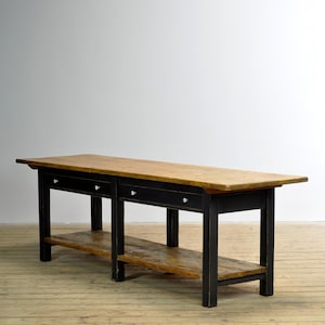 Op de afbeelding: Een lange rechthoekige houten tafel met een natuurlijk lichtbruin houten blad en een onderste plank. Het onderstel en frame zijn zwart geschilderd en de voorzijde is voorzien van twee lades, elk met een kleine, ronde, witte keramische knop.