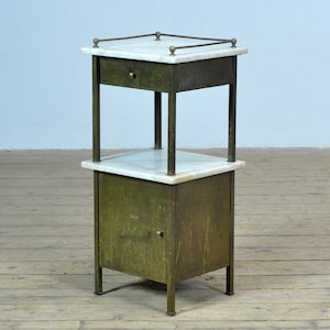 Op de afbeelding: Antieke bijzettafel met een marmeren blad en een lade. De tafel heeft een metalen frame met een verweerde groene afwerking. Het onderste gedeelte heeft een kast met een deur. De tafel wordt ondersteund door vier poten.