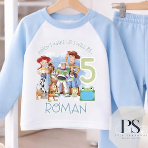 Puede incluir: Un conjunto de sudadera de manga larga y pantalones de chándal a juego en color azul claro y blanco. La sudadera presenta un gráfico de personajes de Toy Story, el texto "WHEN I WAKE UP I WILL BE...", el número "5" y el nombre "ROMAN".