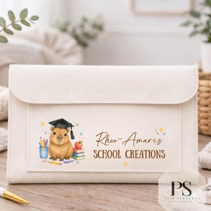 May include: A cream-coloured rectangular pouch with a flap closure, featuring a printed illustration of a capybara wearing a graduation cap, surrounded by school supplies, and the text "Rheo-Amaris SCHOOL CREATIONS" in brown lettering.