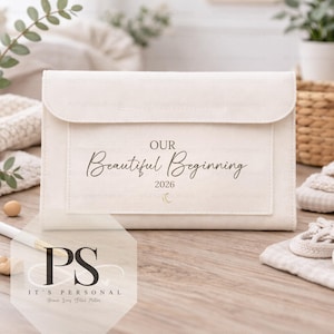 May include: A cream-coloured clutch bag with a flap closure. The front displays the text "OUR Beautiful Beginning 2026" in elegant script, with a small crescent moon. The clutch is on a wooden surface, with a pen and other items in the background.
