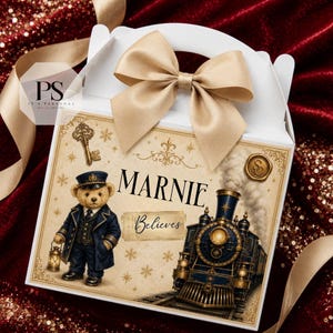 Könnte beinhalten: Eine weiße Geschenkbox mit einer großen goldenen Satinschleife, bedruckt mit einer Illustration eines Teddybären in Schaffneruniform mit Laterne, einer Dampflokomotive, einem antiken Schlüssel und dem Text "MARNIE Believes".