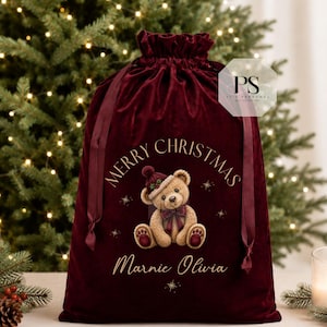 May include: A deep burgundy velvet drawstring gift bag featuring a gold printed Merry Christmas greeting and the name Marnie Olivia. The center graphic depicts a light brown teddy bear wearing a dark red knit hat and a matching plaid bow tie.