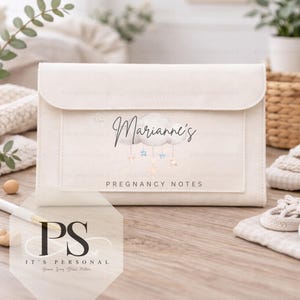 May include: Cream-colored pregnancy notes organizer with a flap closure. The front features the name "Marianne's" above a cloud and star design, with "PREGNANCY NOTES" below. The organizer is on a wooden surface with a pen and other items.