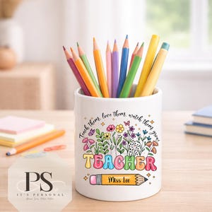 Op de afbeelding: Een witte keramische pennenhouder gevuld met kleurrijke kleurpotloden. De voorzijde is voorzien van een bloemmotief met de tekst "Teach them, love them, watch them grow", het woord "TEACHER" in gekleurde letters en een potloodafbeelding met "Miss Lee".