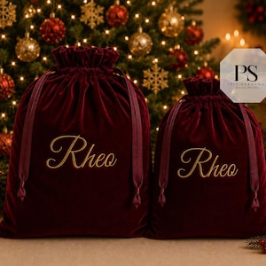May include: Two deep red velvet drawstring gift bags featuring the name Rheo embroidered in gold cursive script on the front. Each bag has matching dark red ribbon ties at the top, creating a gathered closure. The bags are displayed side by side.