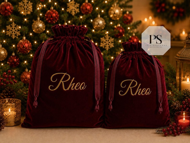 May include: Two deep red velvet drawstring gift bags featuring the name Rheo embroidered in gold cursive script on the front. Each bag has matching dark red ribbon ties at the top, creating a gathered closure. The bags are displayed side by side.