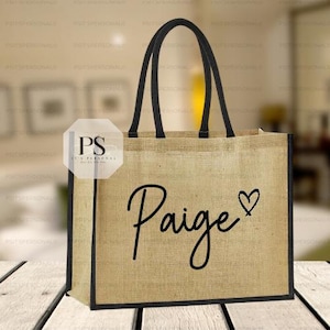 Könnte beinhalten: Eine beigefarbene Jute-Tragetasche mit schwarzen Verzierungen und Griffen. Der Name "Paige" ist in schwarzer Schreibschrift geschrieben, mit einem kleinen Herzsymbol daneben. Die Tasche steht auf einer Holzoberfläche.