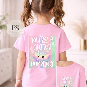 May include: A pink t-shirt featuring the phrase "You're a Glitter Dumpling" and a colourful dumpling graphic. The design includes a rainbow-coloured dumpling with a smiling face, stars, and stripes. A smaller version of the design is on the lower right.