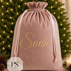May include: A dusty rose velvet drawstring gift bag featuring the name Sadie embroidered in gold script lettering on the front. The bag has a ruffled top edge and matching satin ribbon ties that cinch the opening closed for secure storage.