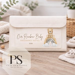 May include: A cream-colored pregnancy notes organizer with a flap closure. The front features the text "Our Rainbow Baby Pregnancy Notes 2026" and a watercolor illustration of a teddy bear on a rainbow. A pen and small wooden beads are on the table.